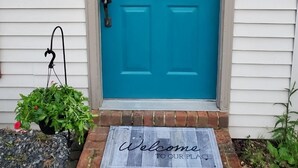 A blue door with a 'WELCOME stay awhile' sign, a doormat with 'Welcome to our place' text, and a hanging planter with greenery.