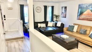A modern living room with a black leather sofa, a brown leather sofa, and a glass coffee table. There is a clock on the wall and a welcome sign above the door.