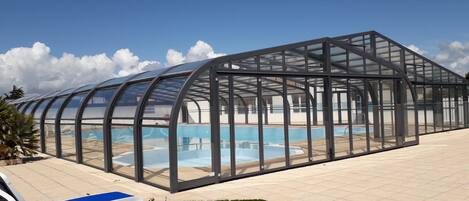 A covered swimming pool with clear glass walls, surrounded by lounge chairs and a well-maintained lawn.