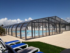 A covered swimming pool with clear glass walls, surrounded by lounge chairs and a well-maintained lawn.