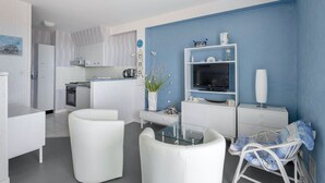 A modern kitchen with white cabinets, a dining area with white chairs, a TV on a white stand, and a blue accent wall. - 50m from the beach, seaview flat for 5 (Pornichet)