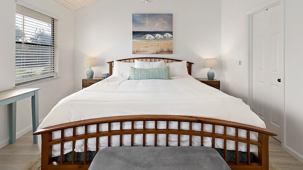 A bedroom with a wooden bed frame, white bedding, two bedside tables with lamps, a ceiling fan, a window with blinds, and a painting of a beach scene.