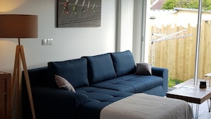 A modern living room with a dark blue sofa, a white throw blanket, and a wooden coffee table. There is a painting on the wall and a lamp on a wooden stand. - Maison Plein Coeur de Ouistreham Riva Bella (Ouistreham)