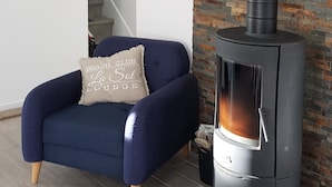 A modern living room with a blue armchair, a stone wall, and a black wood-burning stove. - Maison Plein Coeur de Ouistreham Riva Bella (Ouistreham)