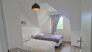 A bedroom with two beds, a sloped ceiling, a window with curtains, and a wooden floor. - Maison Plein Coeur de Ouistreham Riva Bella (Ouistreham)