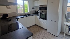 A modern kitchen with white cabinets, a Bosch refrigerator, a built-in oven, and a window providing natural light. - Maison Plein Coeur de Ouistreham Riva Bella (Ouistreham)