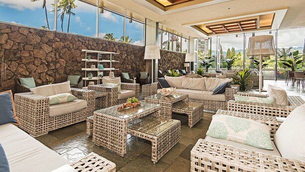 A spacious living area with wicker furniture, a stone wall, and large windows offering a view of palm trees and mountains. - Golf Course & Mountain Views at the Island Colony Amazing Building Amenities (Honolulu)