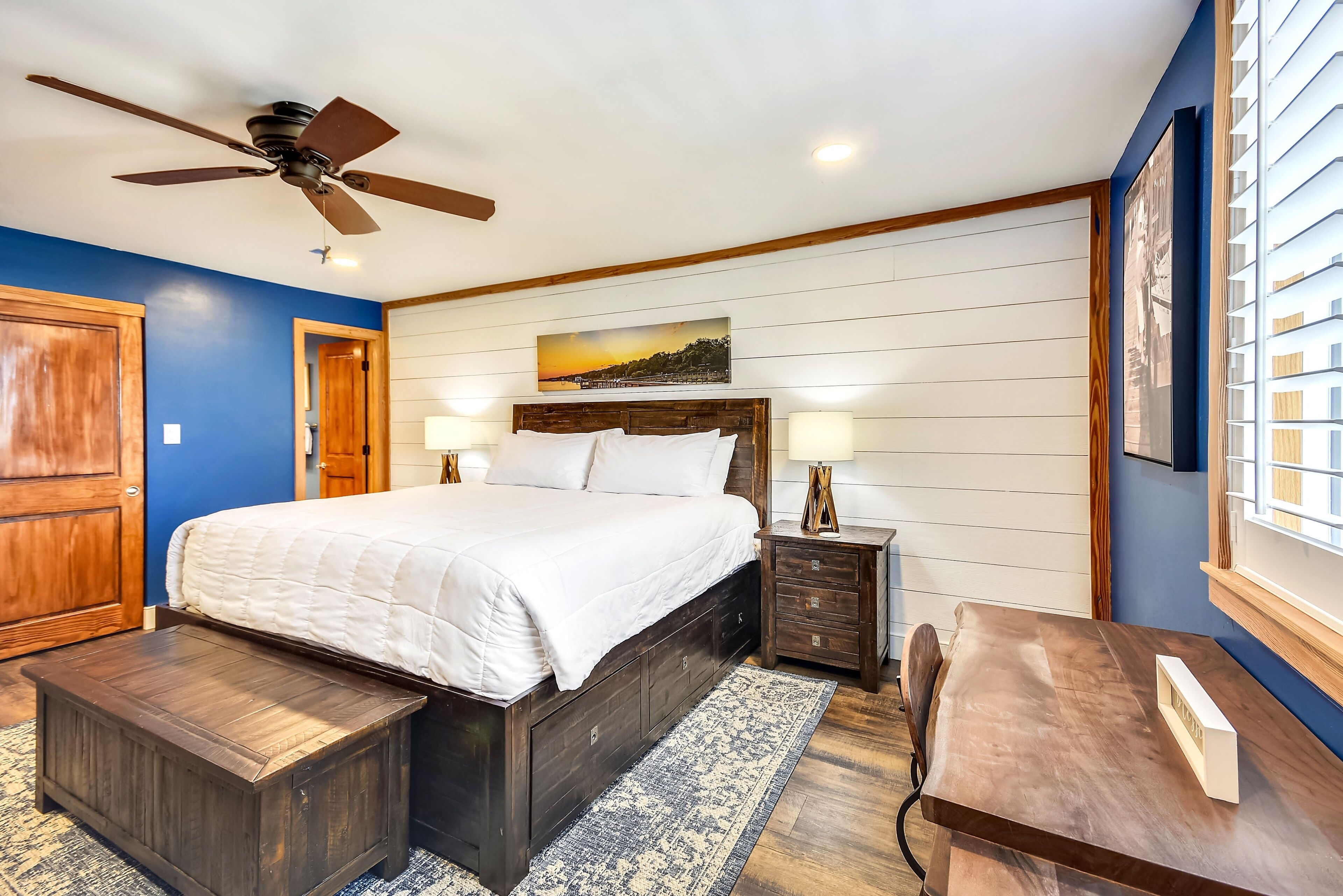 A bedroom with a bed, bedside tables, a desk, a ceiling fan, and a window with blinds.