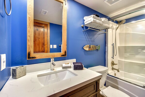 A bathroom with a blue wall, a wooden door, a white sink, a mirror, a towel rack, a shower, and a fish decoration.