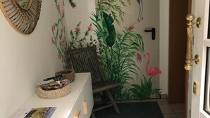 Ein Flur mit einem weißen Konsolentisch, einem Korbstuhl, einem Spiegel und einer Tür mit floralem Wandbild.