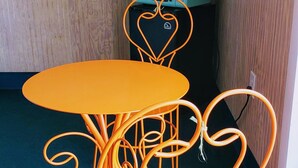 A room with a wooden wall, a television mounted above a green microwave, and a heart-shaped orange metal table and chairs.