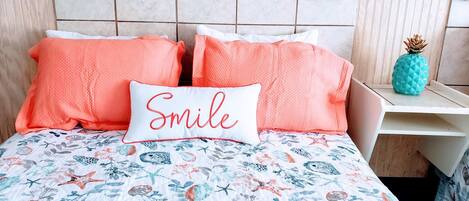 A bed with a floral and sea-themed bedspread, two coral pillows, and a pillow with "Smile" embroidered in red.