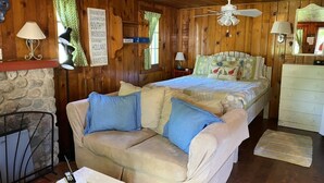A cozy room with a wooden interior, a bed with a floral bedspread, a sofa with blue cushions, a stone fireplace, and a ceiling fan.