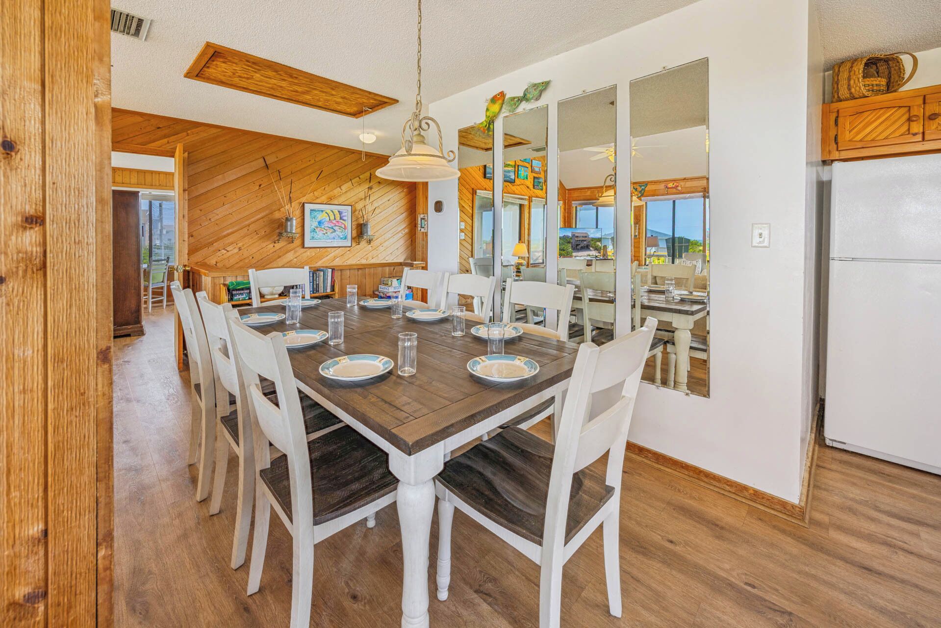 Un coin repas avec une table en bois dressée pour six personnes, des chaises blanches et un grand miroir qui reflète la cuisine.