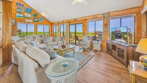 A spacious living room with a large sofa, a glass coffee table, wicker chairs, and a view of the landscape through large windows.