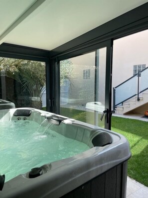 A hot tub in a covered outdoor area with a view of a garden and a staircase leading to a building.