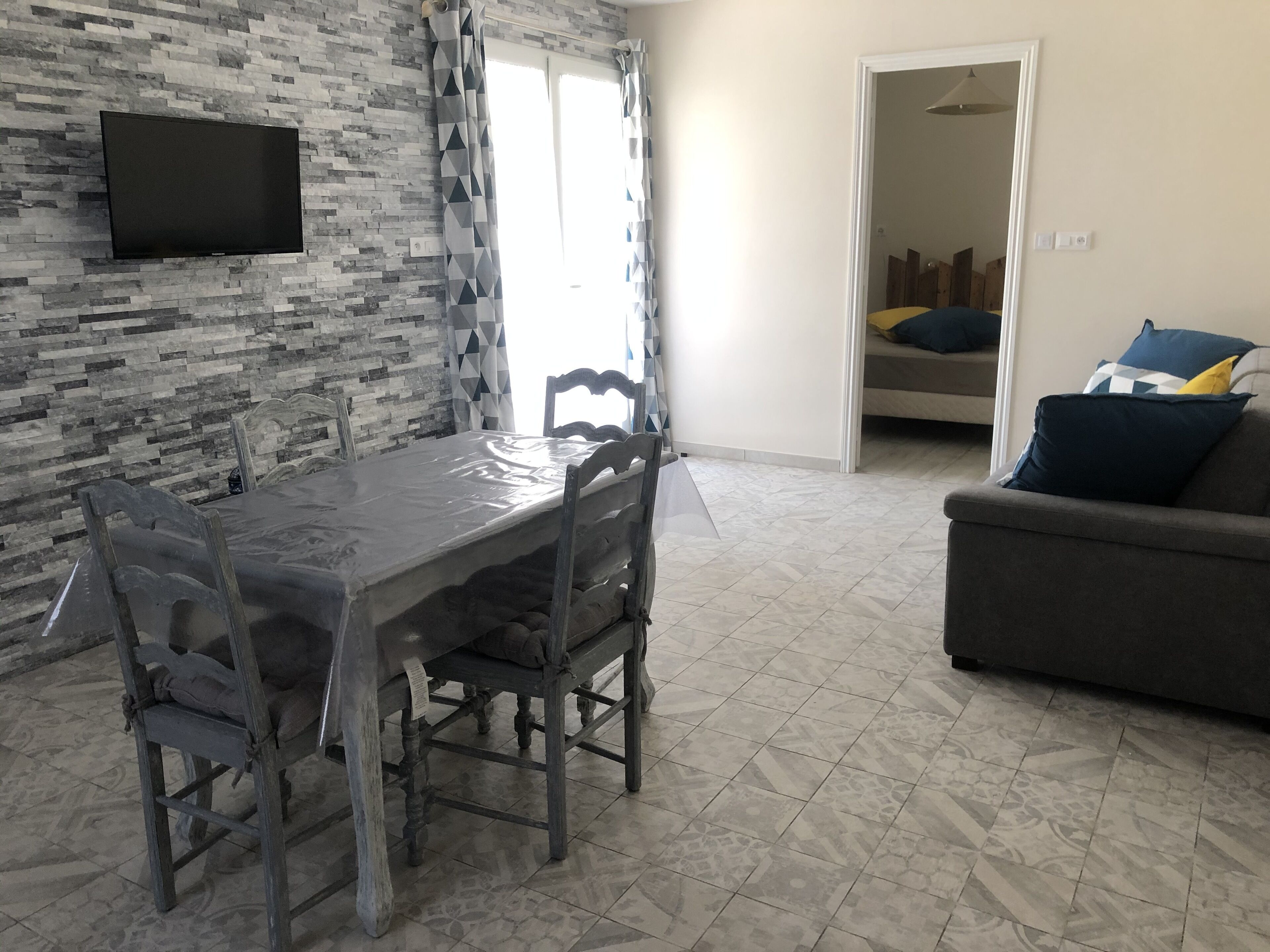 Un coin repas avec une table et des chaises, un téléviseur fixé à un mur en pierre, et un canapé avec des coussins dans l’espace salon.