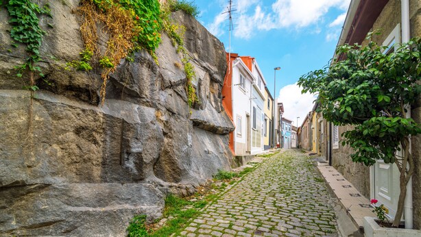 Une rue pavée bordée de bâtiments des deux côtés, un mur rocheux couvert de végétation et un ciel bleu clair.