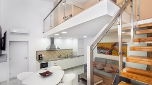 Une cuisine moderne avec des meubles blancs, un four à micro-ondes et une machine à café. On y trouve un coin repas avec une table et des chaises blanches. Un escalier en verre mène à une mezzanine équipée d’un canapé et de coussins.