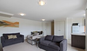 A modern living room with a dark grey sofa, a grey armchair, and a small coffee table. There is a white door and a white cabinet with a microwave on top. - Central one bedroom unit, Thompson Ave Cowes, security entrance, no lift (Cowes)