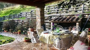 An outdoor patio with stone flooring, white metal chairs, a table set for dining, and a backdrop of stone steps and greenery.