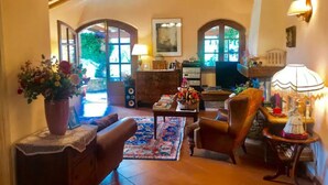 A cozy living room with a sofa, armchairs, a coffee table, and a fireplace. There are two windows with arched tops and a ceiling fan.