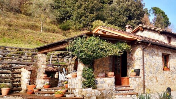 A rustic stone house with a wooden porch, surrounded by potted plants and a hillside backdrop.