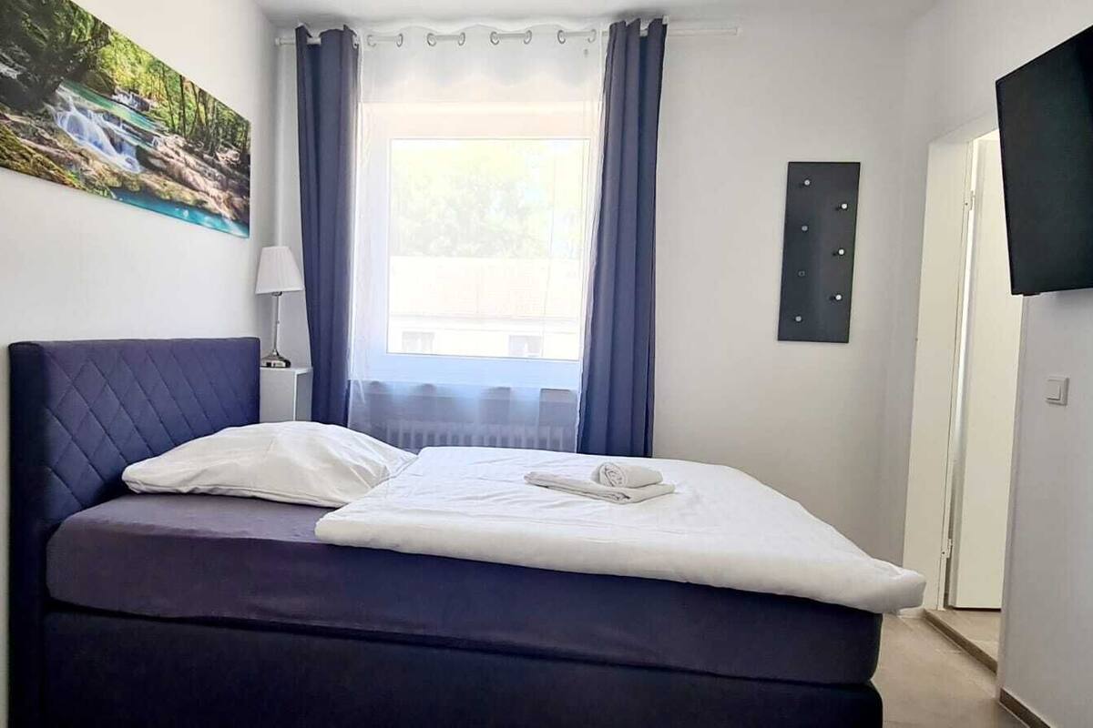 A neatly made bed with a pillow and a folded towel, a window with curtains, a wall-mounted TV, and a painting of a waterfall.