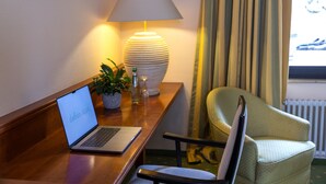 A hotel room with a wooden desk, a laptop, a lamp, a chair, a potted plant, and a window with curtains. - dreams Beckum (Beckum)