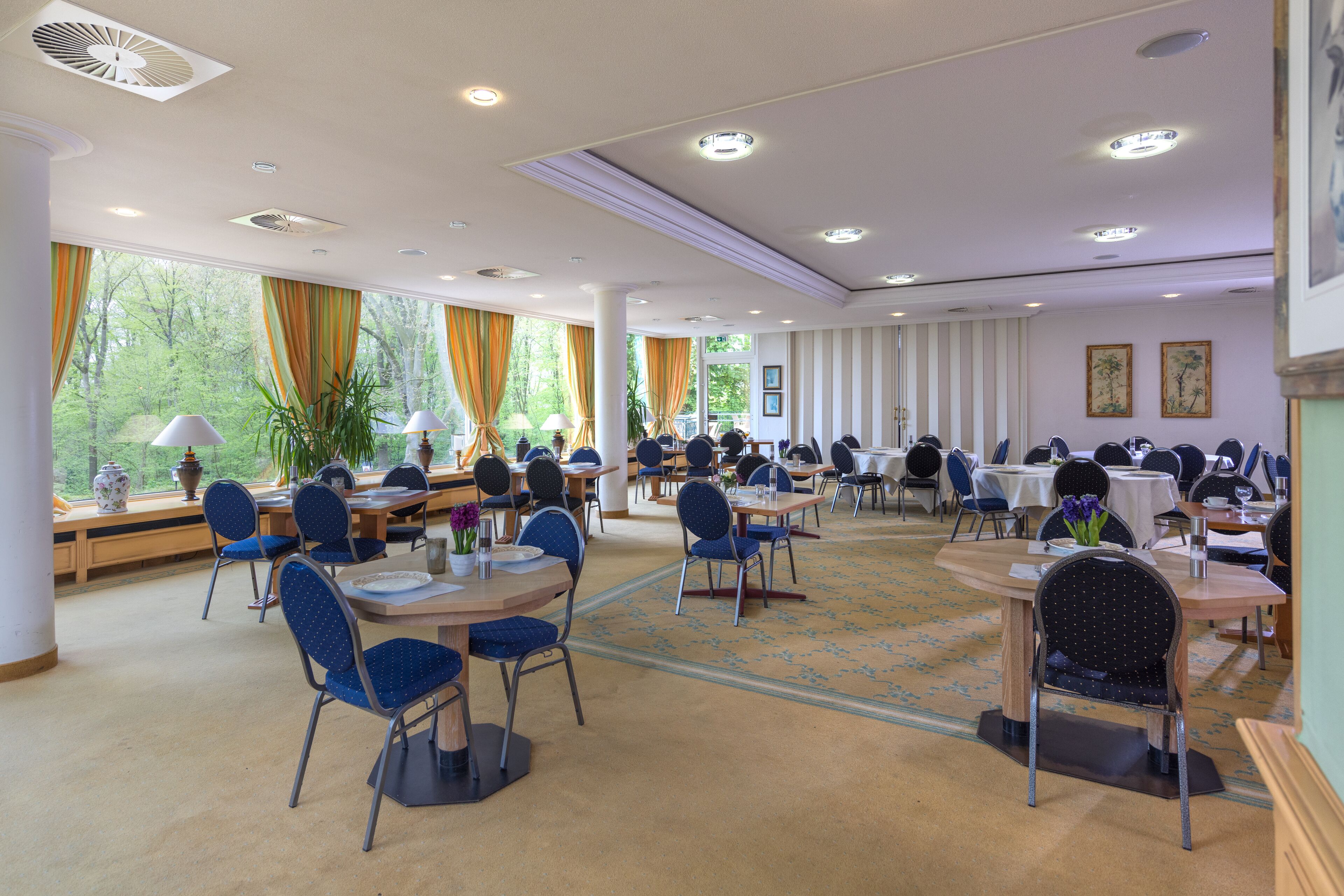 Un comedor amplio con varias mesas y sillas, ventanas grandes con cortinas y vistas a árboles.