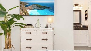 A room with a coastal landscape painting, a white dresser with leather handles, a potted plant, and a lamp.