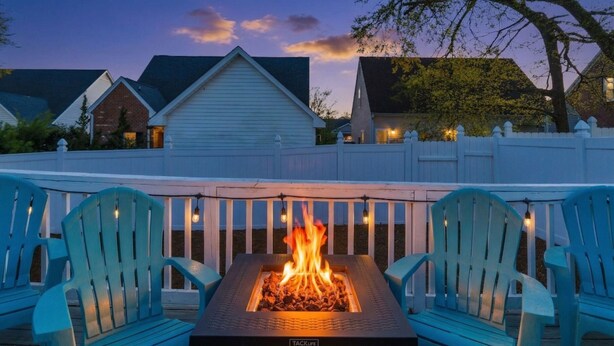 Una terraza con una chimenea, dos sillas Adirondack y una cerca de piquetes blancos.