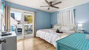 Un dormitorio con cama, ventilador de techo, mesita de noche, lámpara, ventana con cortinas y vistas al exterior.
