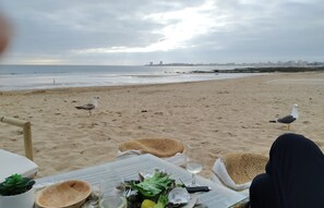Una mesa en la playa con comida, una copa de vino y vistas a la playa y al horizonte de la ciudad.