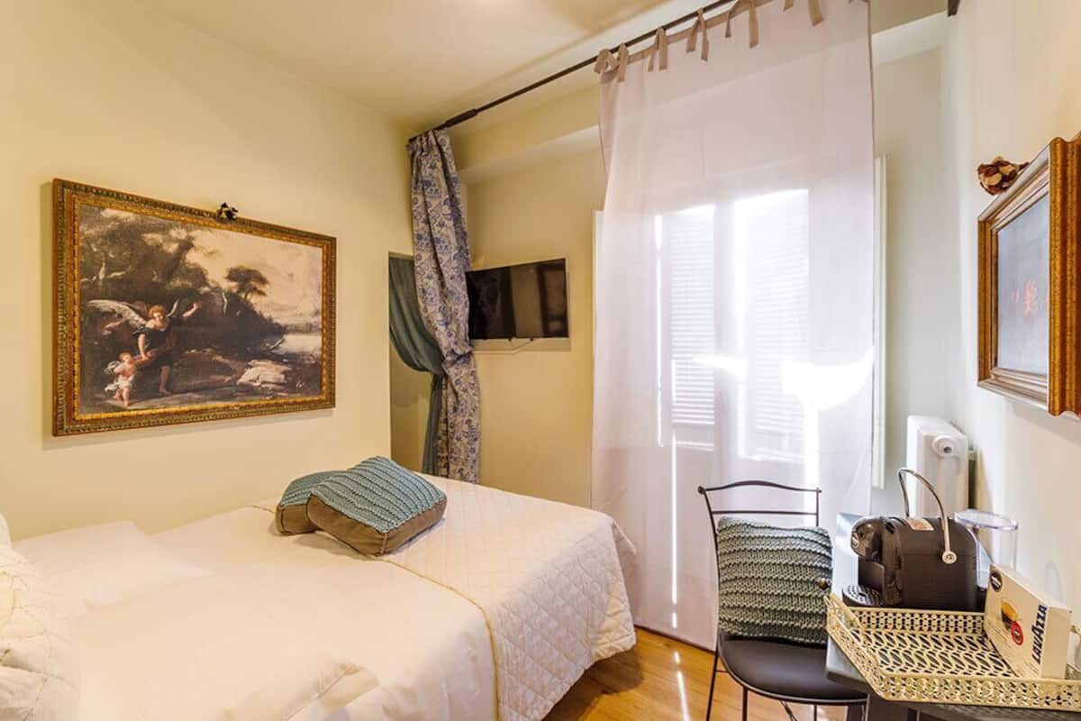 Ein Schlafzimmer mit einem Bett, einem Stuhl, einem Tisch mit Korb, einem Gemälde und einem Fenster mit Vorhängen.