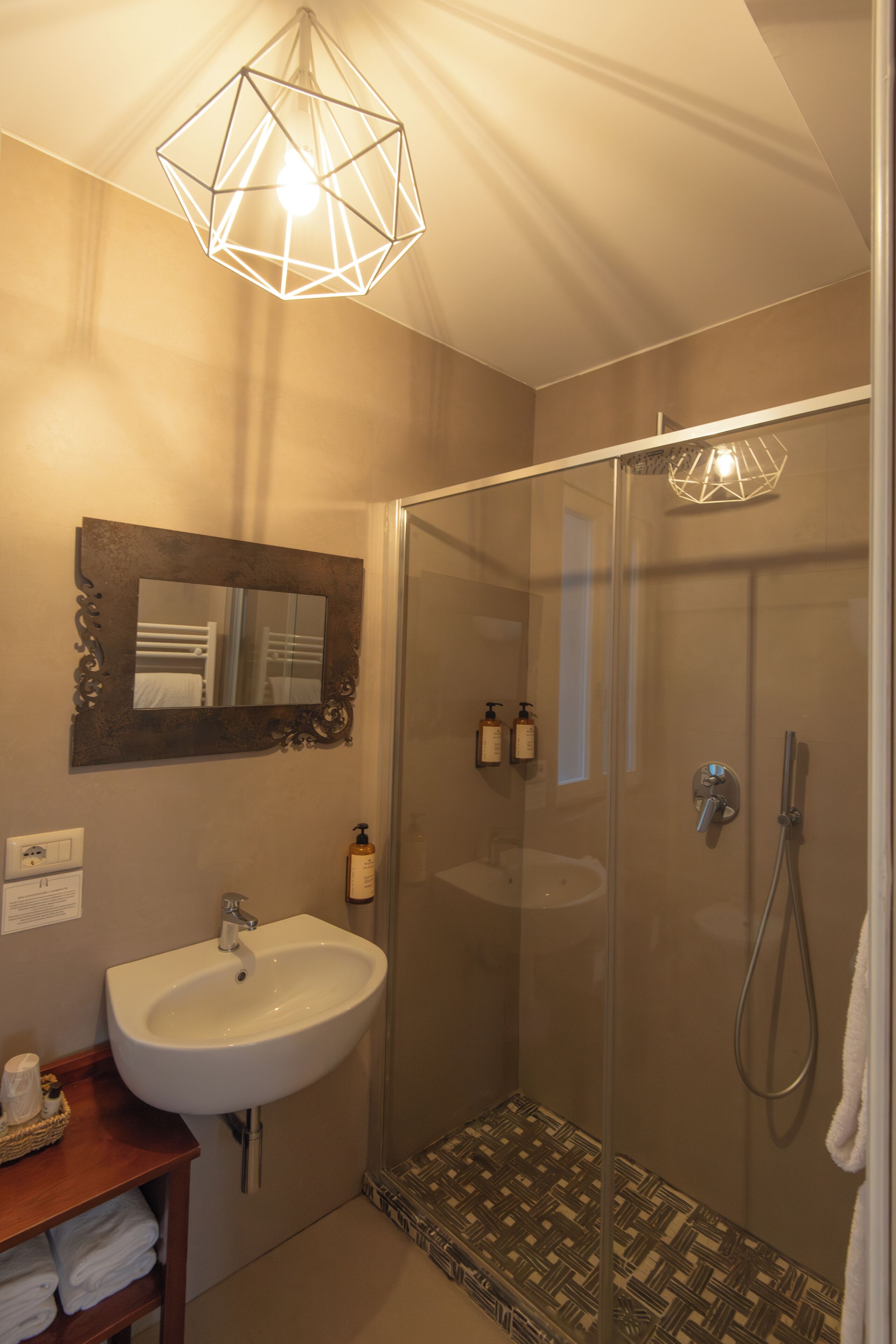 Une salle de bain avec une douche à parois de verre, un lavabo blanc, un miroir et un luminaire au design géométrique.