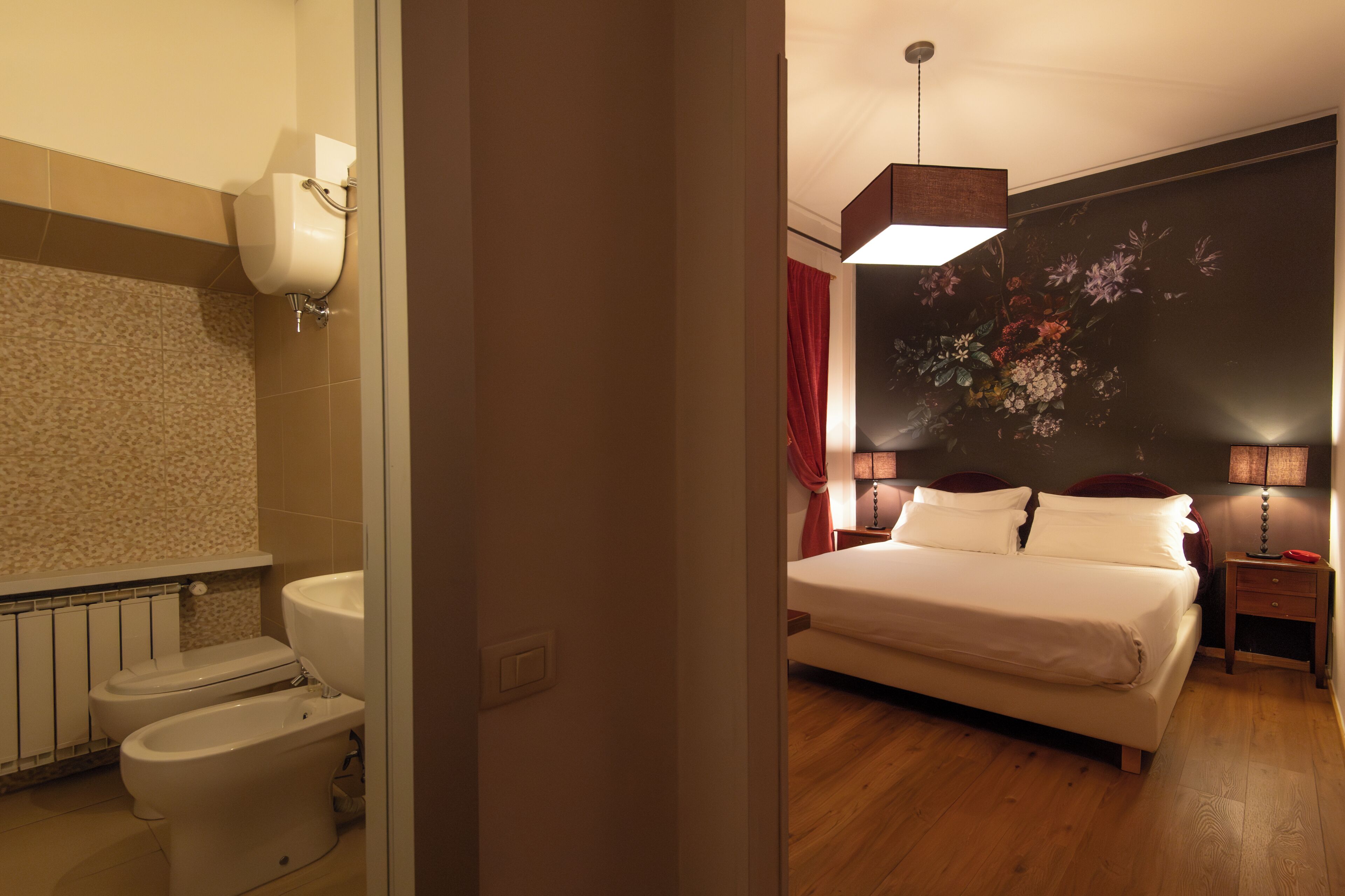 A hotel room with a bathroom, a bed, a nightstand, and a wall with a floral painting.