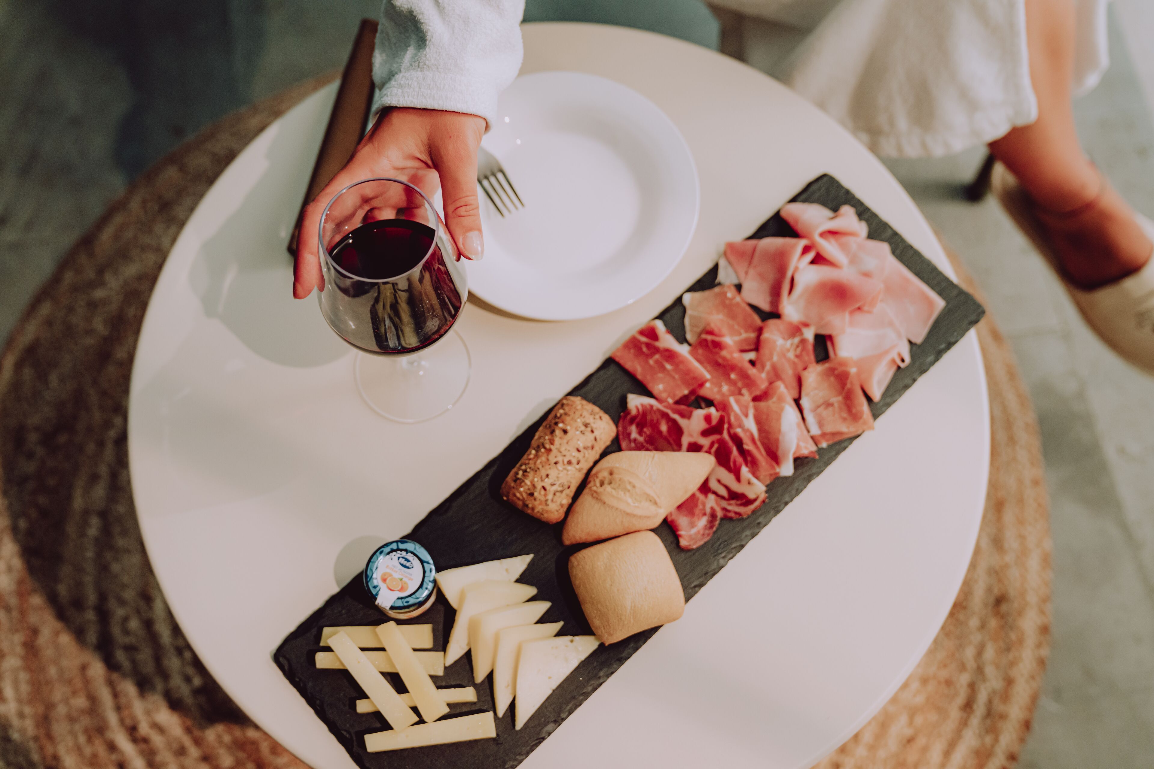 A round table with a white plate, a glass of red wine, a cheese board with various cheeses, and a selection of cured meats.