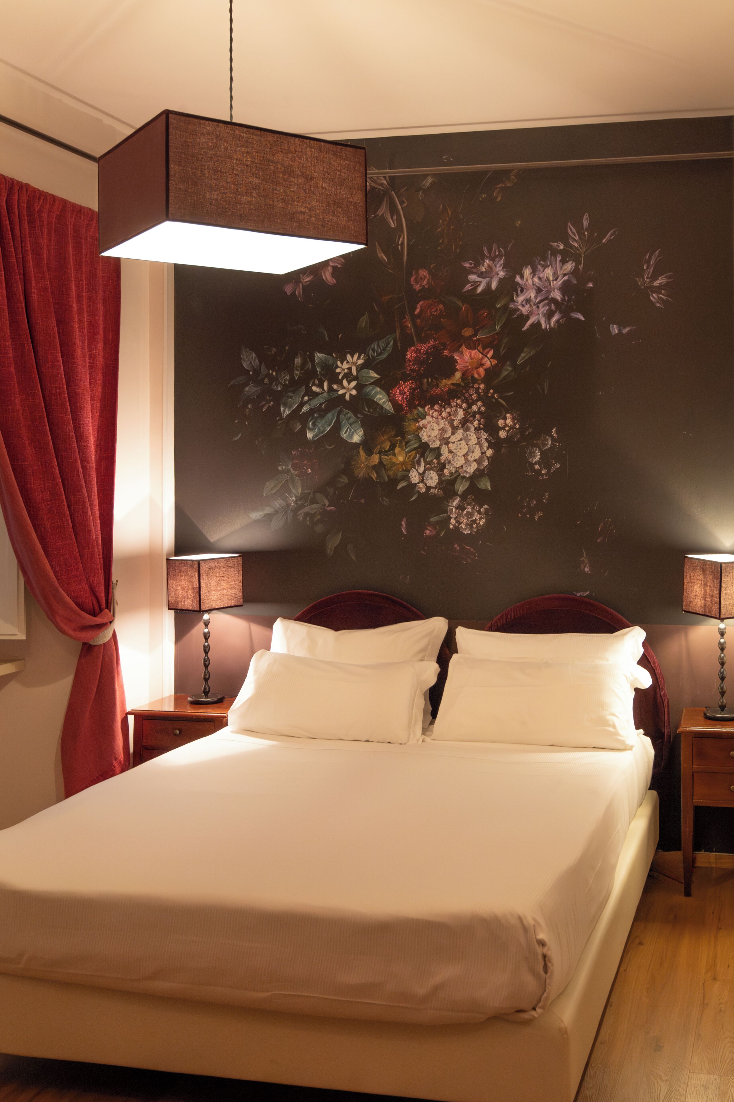 Een keurig opgemaakt bed met witte lakens, twee nachtkastjes met lampen, een rood gordijn en een bloemen behang.