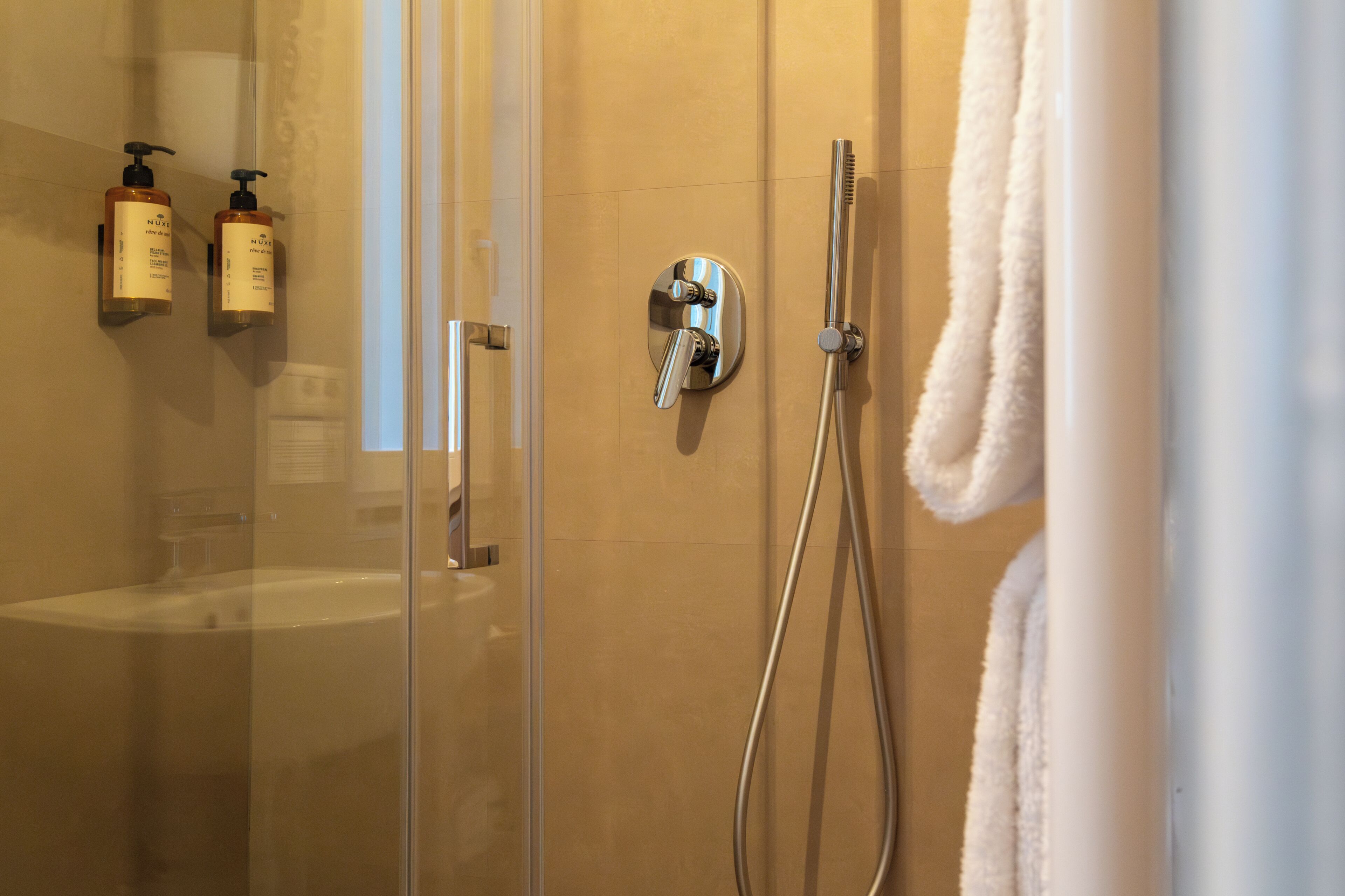 A glass-enclosed shower with a handheld showerhead, a wall-mounted faucet, and two bottles of shampoo on the wall.