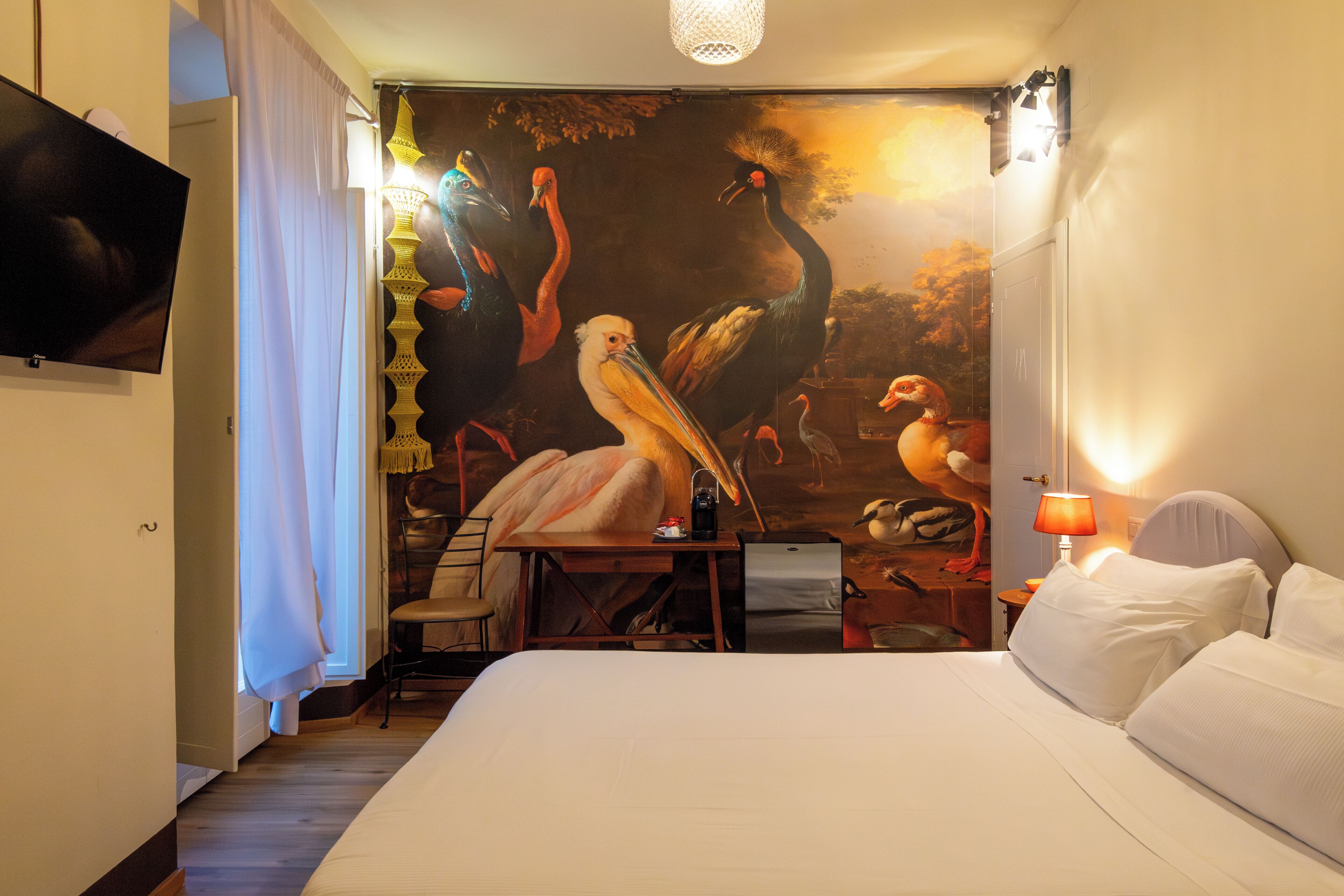 Et hotelværelse med et stort mural af fugle, en seng, et skrivebord med stol, et fjernsyn og en lampe.