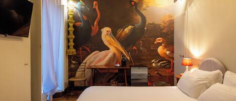 A hotel room with a large mural of birds, a bed, a desk with a chair, a television, and a lamp.