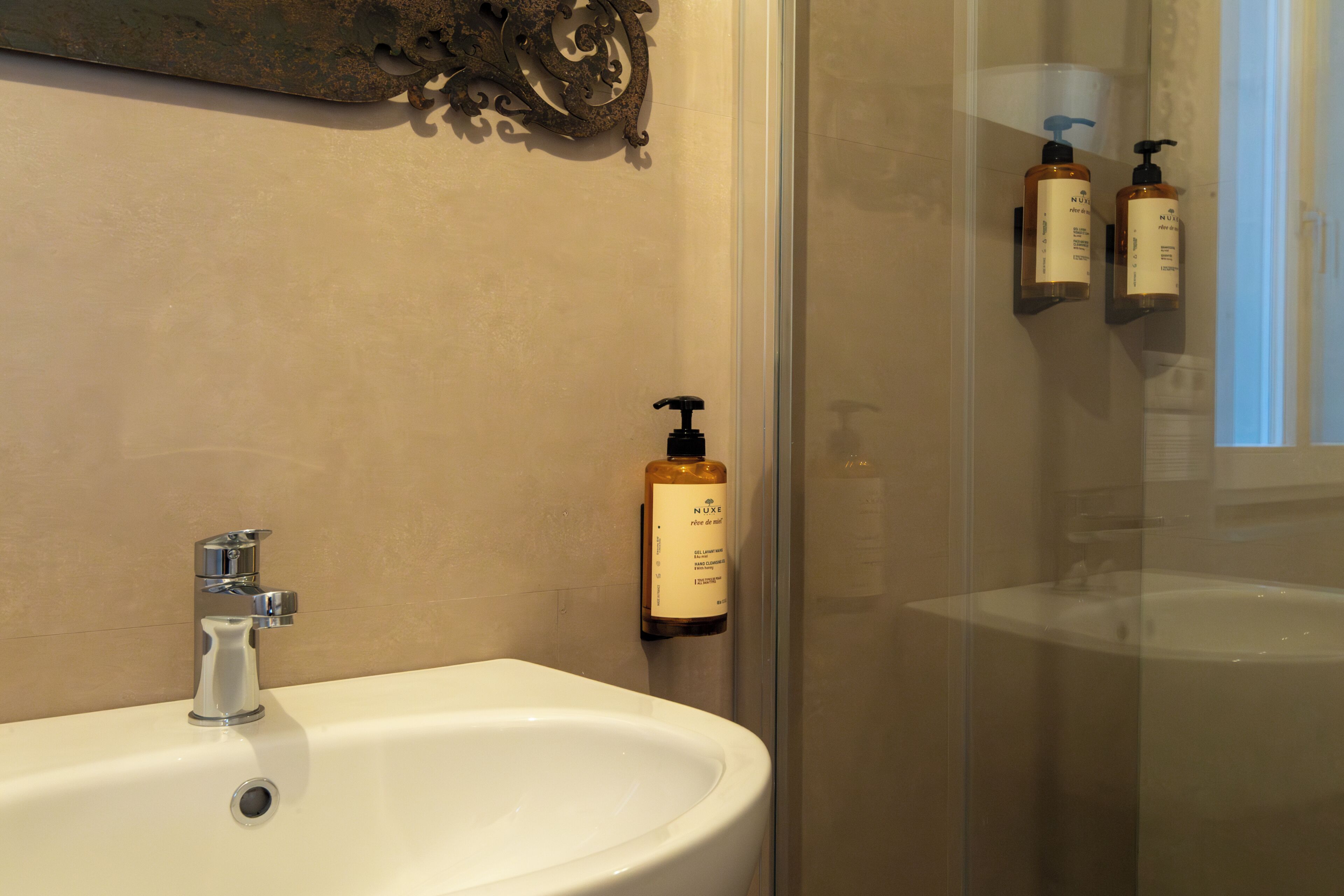 A bathroom with a white sink, a glass shower enclosure, and two Nuxe hand soap dispensers mounted on the wall.