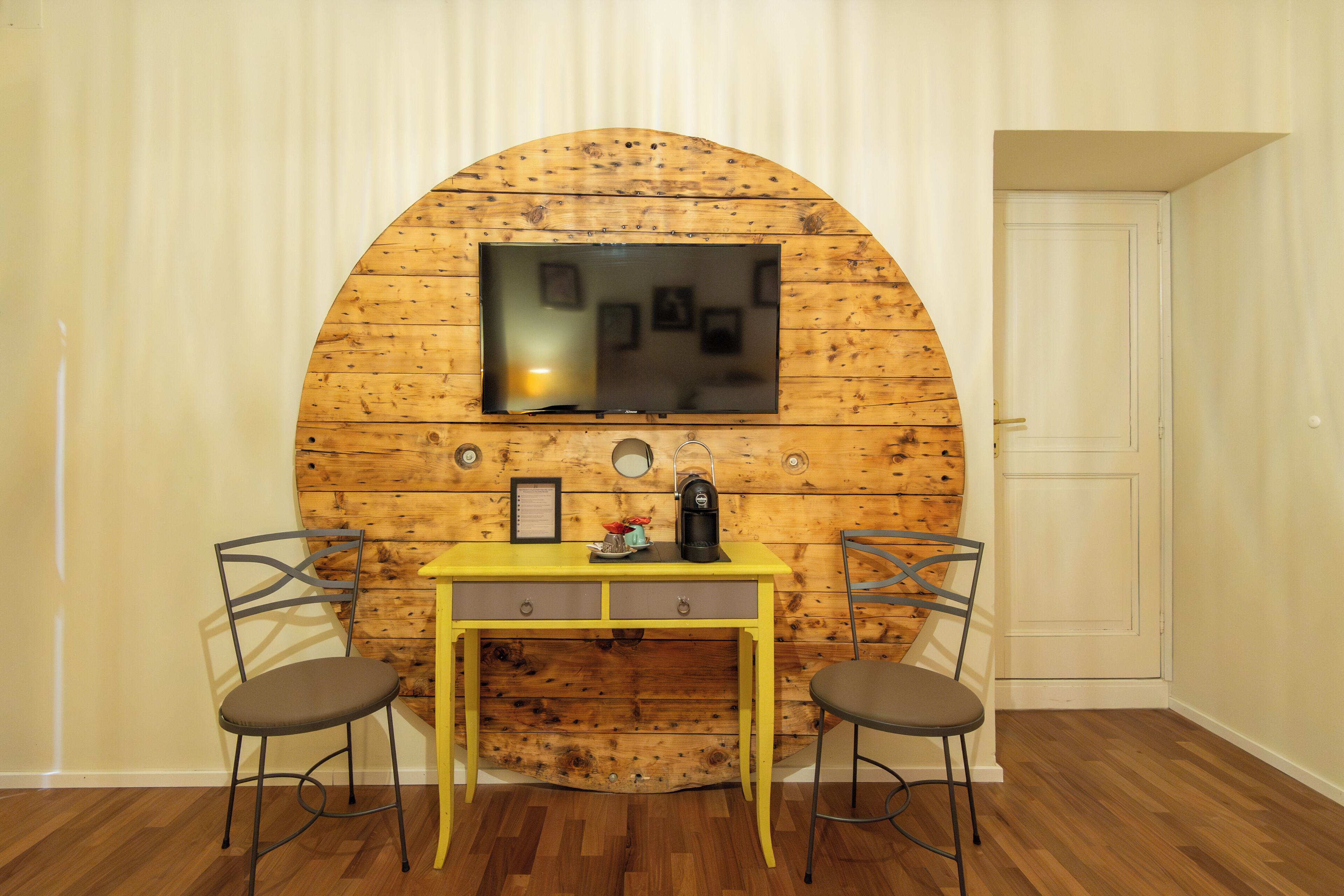 A room with a wooden wall, a TV mounted above a yellow desk, two chairs, and a door.