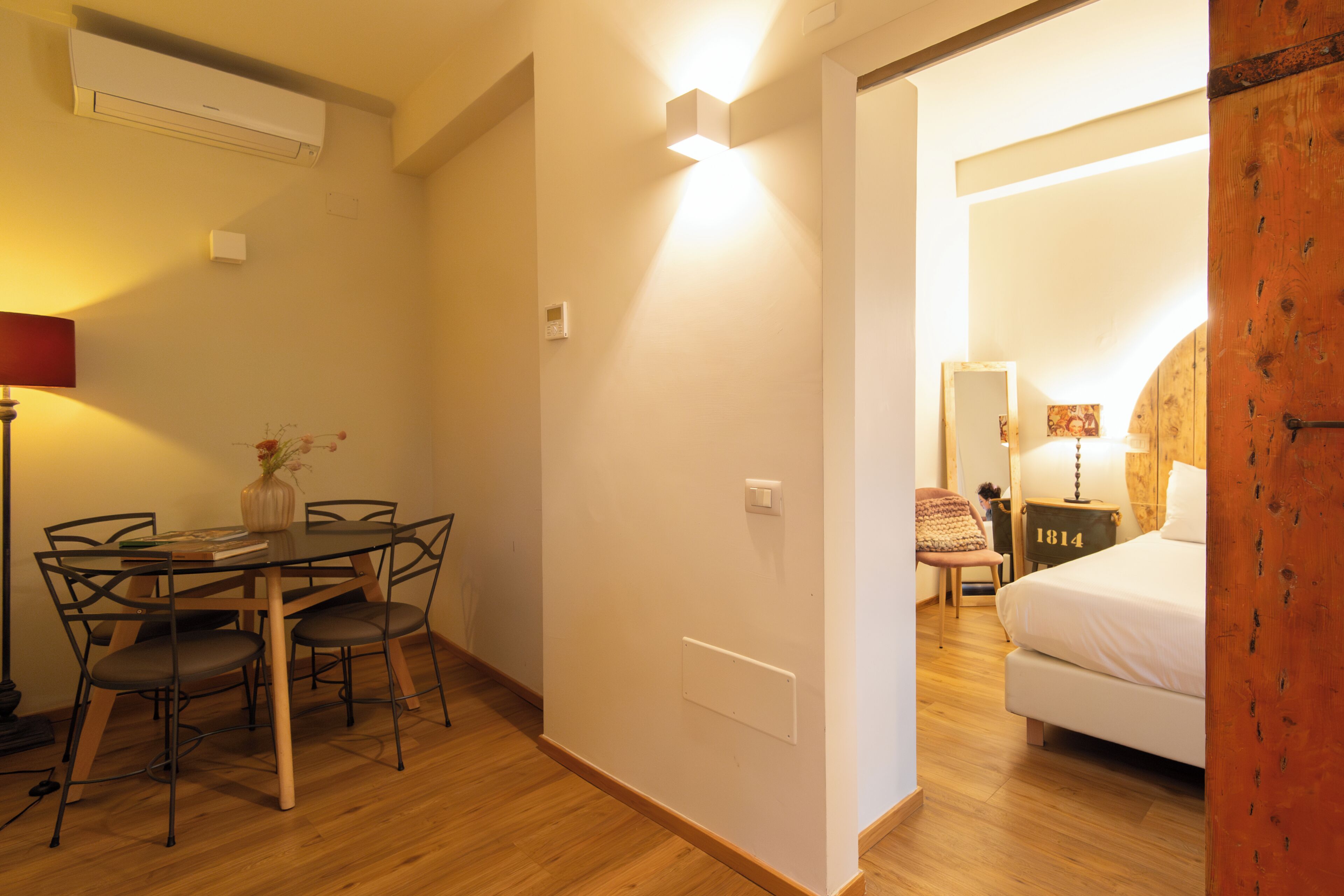 Una stanza con pavimento in legno, un tavolo da pranzo con sedie, un letto e un comodino con una lampada.