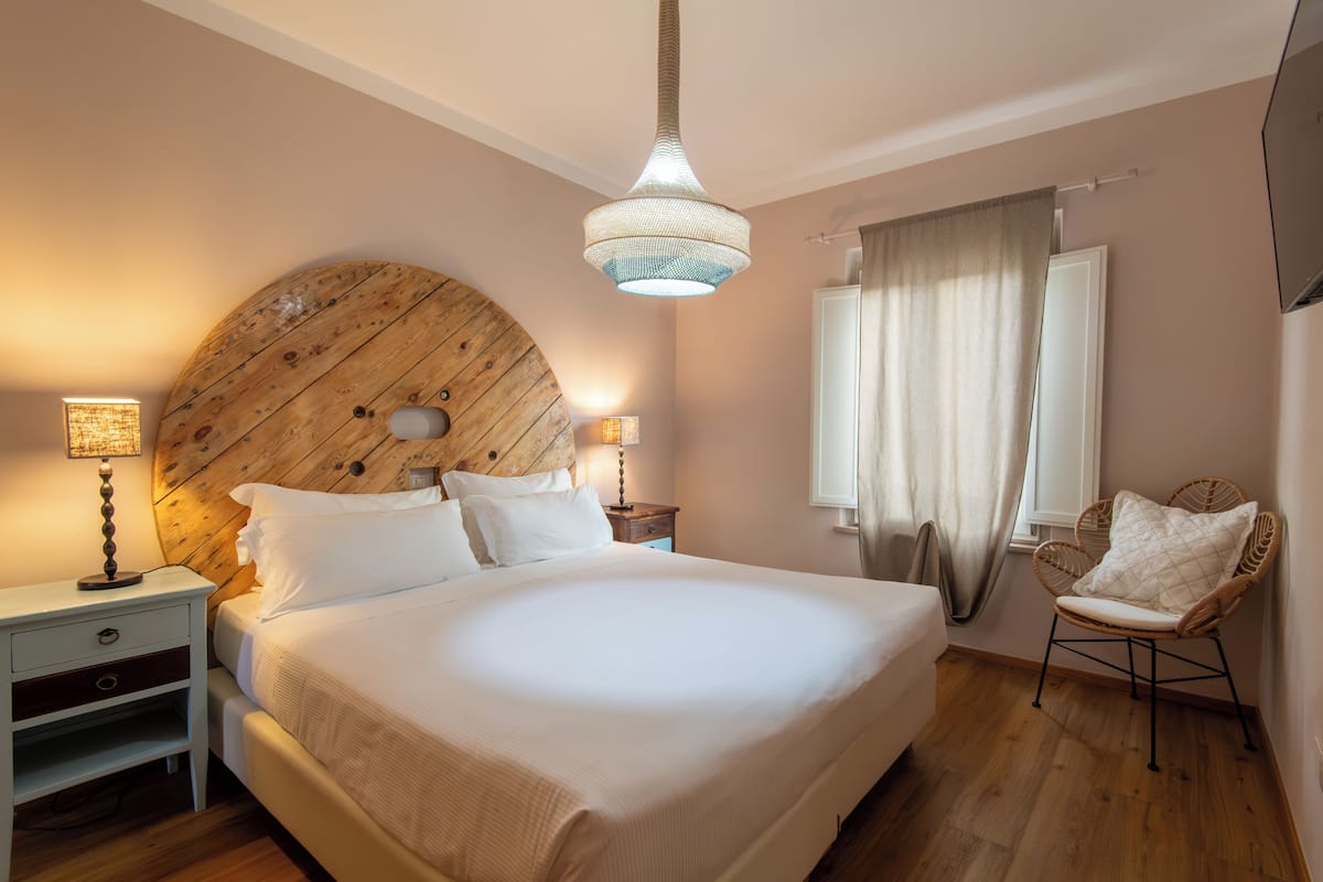 Ein Schlafzimmer mit einem großen Bett, einem Holzkopfende, Nachttischlampen, einem Stuhl und einem Fenster mit Vorhängen.