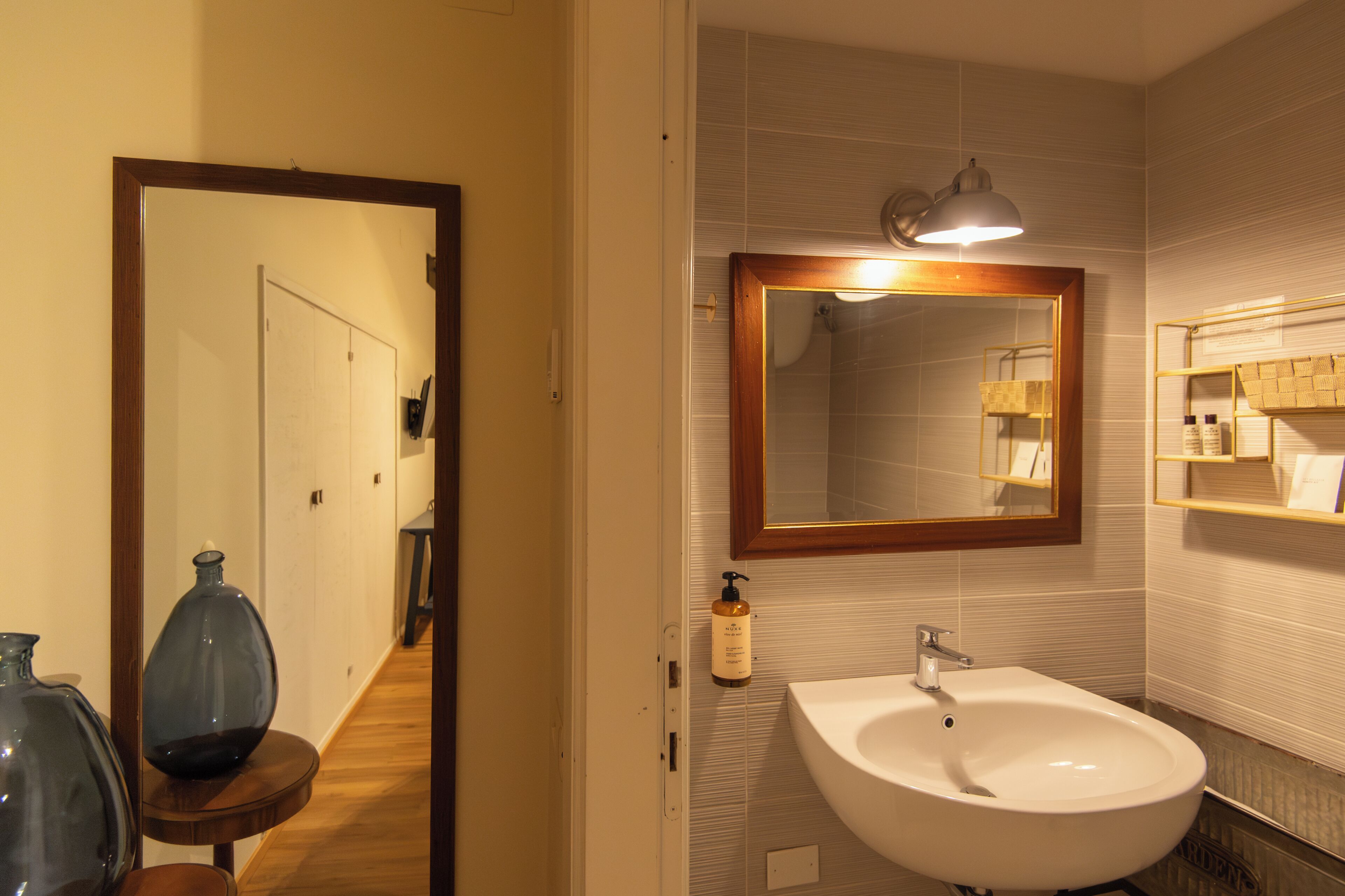 Un baño con lavamanos blanco, un espejo con marco de madera y un estante con artículos de aseo.