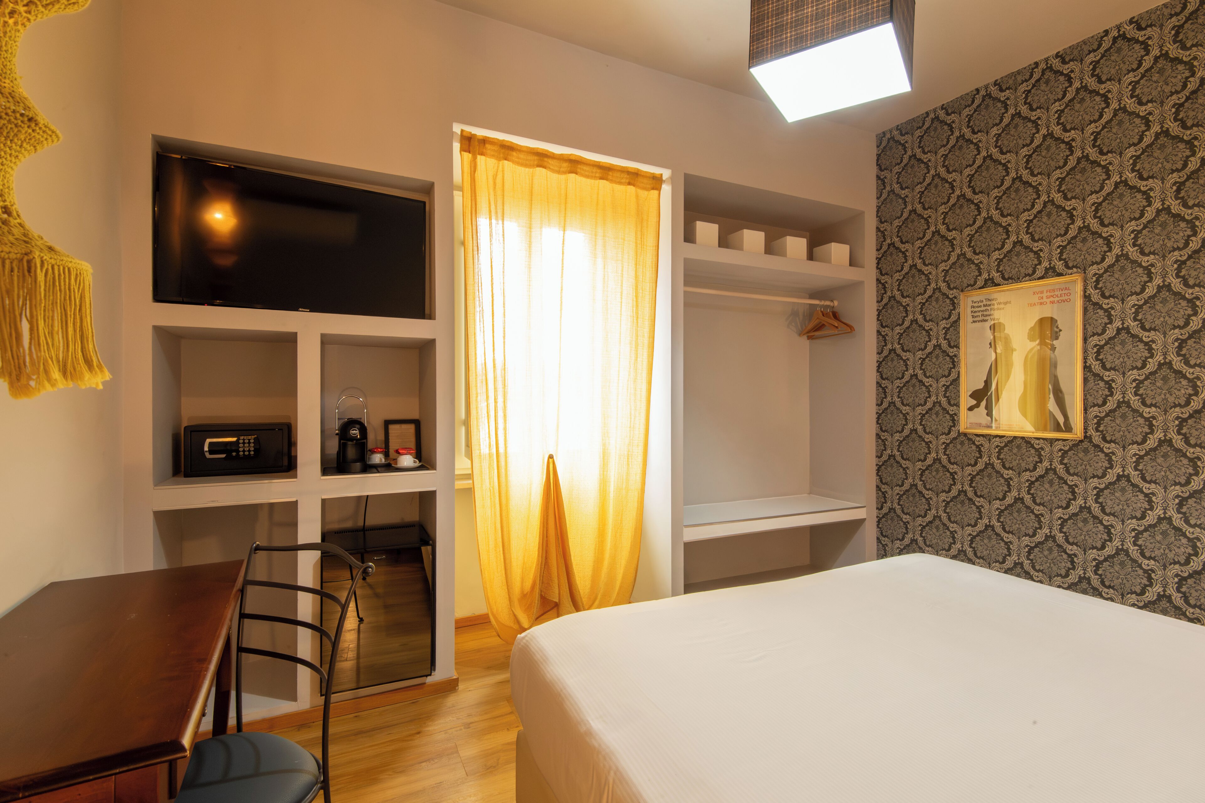 Une chambre d’hôtel comprenant un lit, un bureau, une chaise, une télévision, un four à micro-ondes, une machine à café, une étagère, une armoire et un mur décoré de motifs.