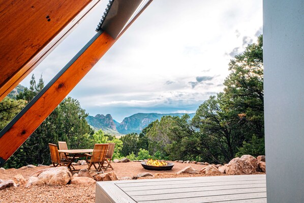 An outdoor patio with a wooden table and chairs, a fire pit, and a view of mountains and trees.