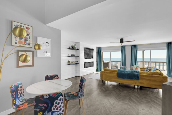 Una sala de estar moderna con un sofá amarillo, una mesa de comedor redonda con sillas coloridas y un amplio ventanal con cortinas.